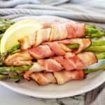 Bacon wrapped asparagus on white plate with lemon wedges.
