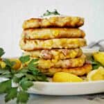Fritters stacked in a pile on white plate with parsley and lemon wedges.