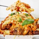 Spoon holding ziti pasta over white baking dish of the same.