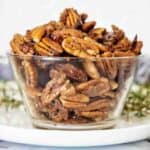 Glass bowl of candied nuts on white plate with rosemary leaves behind it.