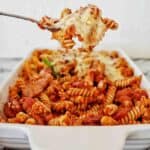 Spoon holding baked chorizo pasta above white baking dish of the same.