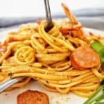 Fork being twirled in pasta on white plate with slices of chorizo and prawns.