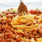 Fork being twirled in fettuccine bolognese on white plate