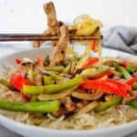 Chopsticks holding garlic pepper beef above white bowl of the same with rice.