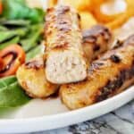 Sausage with the end cut off on white plate with onion rings and salad.
