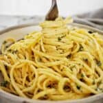 Fork being twirled in bowl of alfredo with linguine with grey tea towel behind.