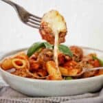 Fork holding meatball with cheese dripping from it over grey bowl of pasta bake.