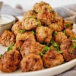 Meatballs piled high on white plate with small bowl of sauce in the background.