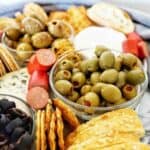 Olives in a glass bowl on a plate with crackers and cheese.