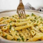 Fork being pushed into penne alfredo in grey bowl.