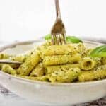 Fork being pushed into pesto rigatoni in grey bowl.