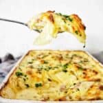 Spoon holding potato bake above white baking dish of the same.
