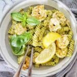 Pasta with pesto and prawns in grey bowl with lemon wedges and basil leaves.