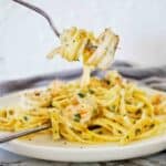 Fork holding prawn fettuccine over white plate of the same.