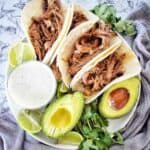Soft shell tacos with pulled beef on white plate with avocado, cilantro and sauce.