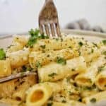 Fork being pushed into rigatoni alfredo in grey bowl.