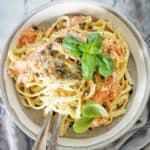 Salmon fettuccine in grey and white bowl with utensils and basil leaves.