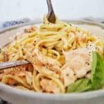 Fork being twirled in salmon linguine alfredo in grey bowl with basil leaves.