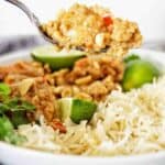 Spoon holding prawns over a white bowl of the same with rice and lime wedges.