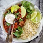 Chilli con carne with rice in white bowl with lime wedges and cilantro.