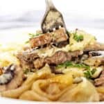 Fork being pushed into cooked beef with onion and mushrooms on a white plate.