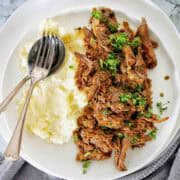 Cooked pulled beef on white plate garnished with parsley and served with mashed potato.