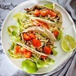 Beef tacos on white plate with cheese and lime wedges.