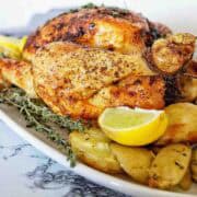 Roast chicken with thyme on white plate with potatoes and lemon wedges.