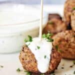 Beef meatball with toothpick through it on white plate with sauce bowl in background.