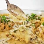 Spoon pouring cream sauce over chicken with mushrooms in white pan.