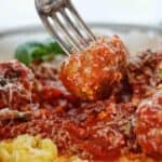 Fork holding meatball above bowl of marinara sauce and polenta.